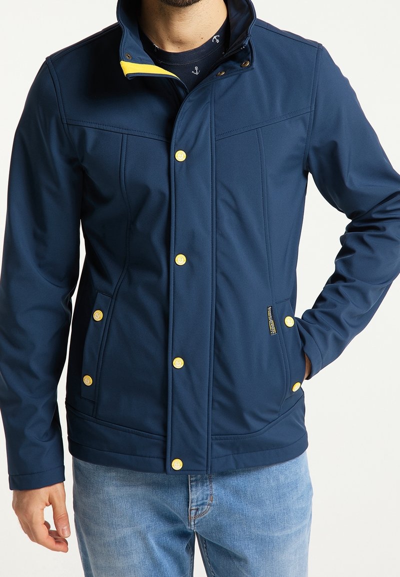 Marineblå softshelljacka med en strukturerad krage, gula accenter och guldknappar. Har sidofickor och en skräddarsydd passform.