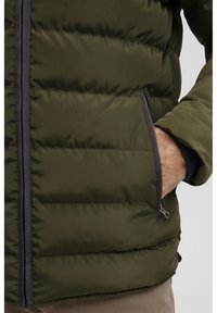 Oliven grønn dunjakke med kviltet design, grå glidelåsdetaljer og sidelommer. Stoffet ser glatt og lett ut.
