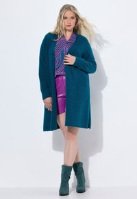 Teal kofta med avslappnad passform, långa ärmar och fickor i lappar, som är kombinerad med en lila mönstrad topp och rosa fusk läder shorts.