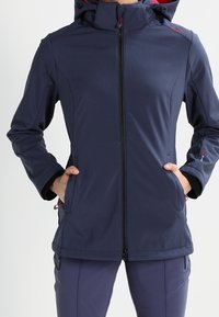 Woman wearing a navy blue zip-up hooded jacket with hands in front pockets, paired with matching navy pants.