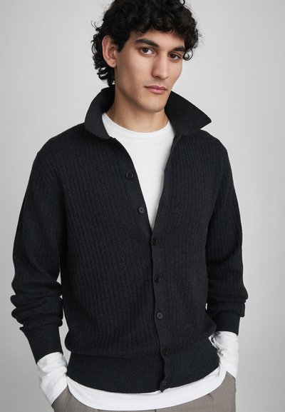 Giovane uomo con capelli ricci e scuri che indossa un maglione nero a costine con bottoni sopra una camicia bianca a maniche lunghe, mani in tasca, sfondo neutro.