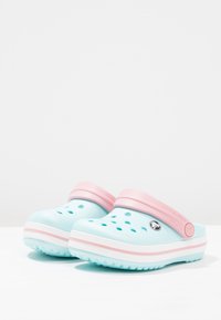 Světlomodré crocsy s růžovým popruhem, obsahující ventilační otvory, texturovanou podrážku a bílý pruh podél základny.