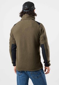Olivgrüne Fleece-Jacke mit schwarzen Panel-Details, hohem Kragen, strukturiertem Stoff und schwarzen langen Ärmeln. Rückansicht einer Person, die Jeans trägt.