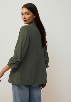 Donna con capelli lunghi e scuri che si volta indietro, indossa un blazer verde oliva con le maniche arrotolate e jeans azzurri su uno sfondo semplice.