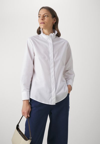 Weiße Baumwollbluse mit einem Rüschensaum, langen Ärmeln und Knopfverschluss, kombiniert mit dunkelblauen Jeans und einer beigen Handtasche.