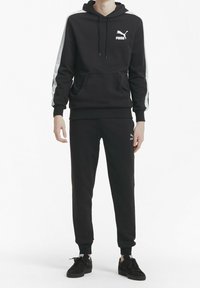 Svart huvtröja med vit Puma-logotyp, har frontficka och snörning. Matchas med svarta byxor och svarta sneakers.