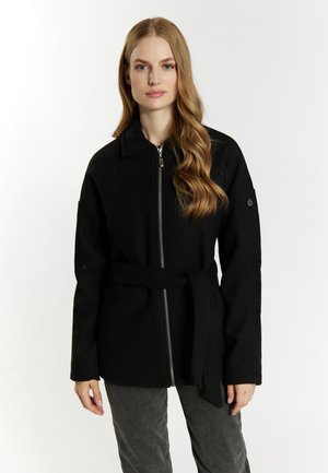 Woman with long wavy hair wears a black zip-up jacket with a belt, standing against a plain light background.