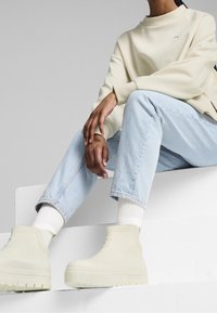 Bottes montantes beiges claires Puma en caoutchouc texturé avec une semelle épaisse et un détail de logo, associées à un jean en denim bleu clair et des chaussettes blanches à côtes.