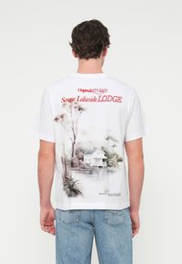 Λευκό βαμβακερό T-shirt με γραφικό τύπωμα στην πλάτη που απεικονίζει μια γαλήνια λίμνη και κείμενο σε κόκκινο χρώμα. Στρογγυλή λαιμόκοψη και κοντά μανίκια.