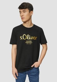 Black cotton t-shirt featuring gold lettering "s.Oliver" and "ELEVATED ESSENTIALS SINCE 1969" on the front, round neckline, short sleeves.