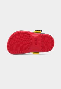 Κόκκινη υφή σόλας παιδικού clog Crocs με ενδείξεις μεγέθους, ορατά μαύρα και κίτρινα επάνω λουράκια σε λευκό φόντο.
