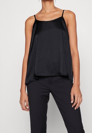 Femme portant un haut caraco en satin noir sans manches associé à un pantalon noir sur mesure, sur un fond uni.