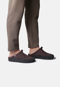 Bruna slip-on skor med en vadderad ovandel och grå filtaccent, med rundad tå och en svart gummisula, visade bäras med grå byxor.