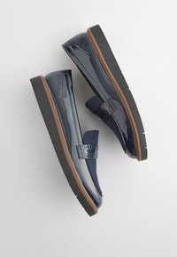 Paire de mocassins en cuir bleu foncé brillant avec des détails brogue, une languette en tissu bleu marine, une bordure marron et des semelles en caoutchouc noir.