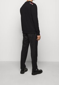 Sudadera negra con puños acanalados, cuello redondo y logo rojo contrastante en la parte trasera. Combinada con pantalones negros ajustados y zapatos negros.