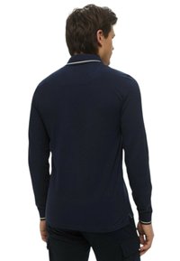 Uomo con capelli corti e scuri che indossa una polo a maniche lunghe blu navy con bordi bianchi su colletto e polsini, visto di spalle.