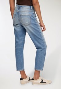 Jeans blu chiaro a vita alta con una vestibilità rilassata, presentano dettagli strappati e gambe dritte, abbinati a sneaker beige e nere.