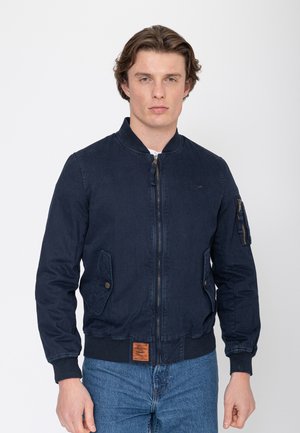 Homme portant une veste bomber bleu marine avec poches avant et fermeture éclair, associée à un jean bleu sur un fond clair uni.