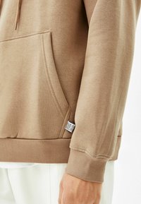 Brązowa bluza z kapturem z przednią kieszenią, ściągaczami na mankietach i u dołu. Posiada materiałową metkę po boku. Gładka faktura, casualowy design.