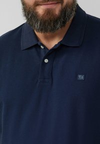 Granatowa koszulka polo wykonana z teksturowanej tkaniny, z klasycznym kołnierzykiem i dwoma guzikami, z małym kwadratowym logo po lewej stronie klatki piersiowej.