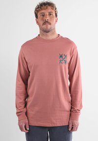 T-shirt en coton à manches longues, rose, avec un petit motif floral bleu sur la poitrine gauche. Texture douce et coupe décontractée.