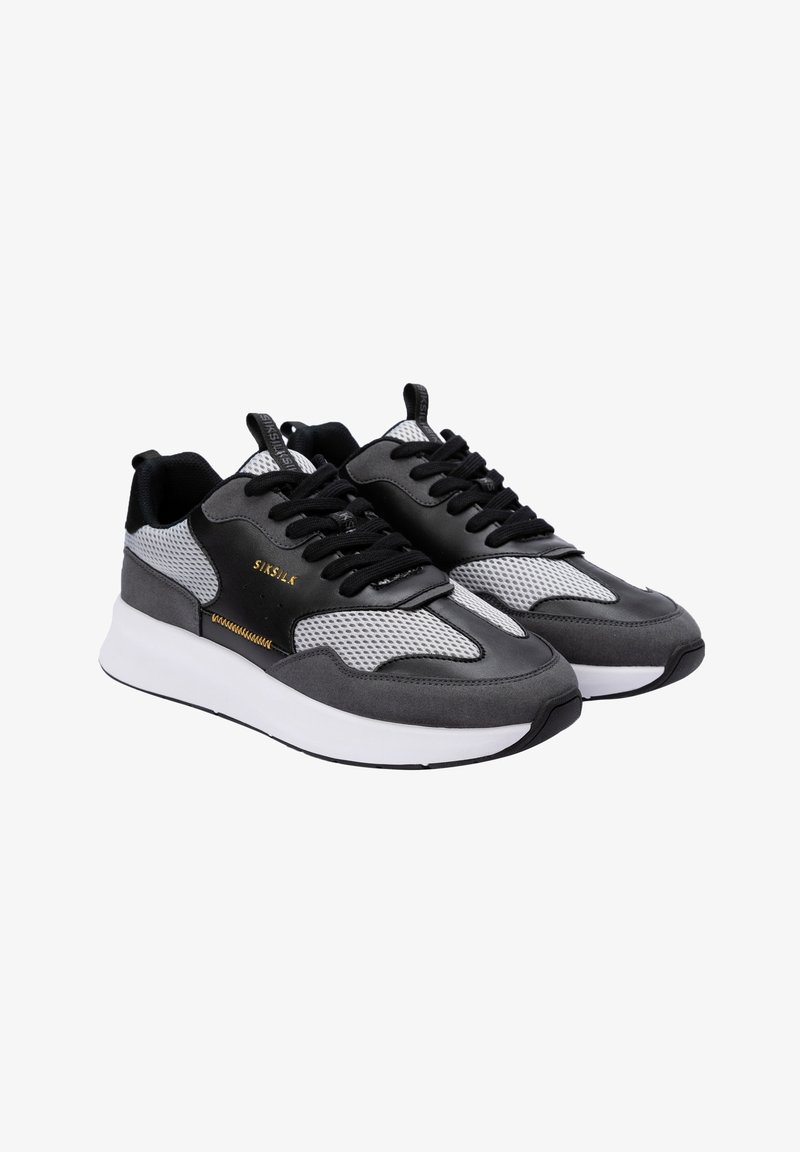 Chaussures de sport noires et grises avec des panneaux en mesh, semelle blanche épaisse, bout rond, lacets noirs et détail de logo doré sur le côté.