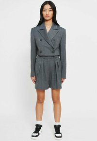 Grijze, dubbelzijdige blazer met schoudervullingen, gecombineerd met bijpassende geplooide short. Heeft een gestructureerde stof en zwarte knopen, gestyled met sneakers.