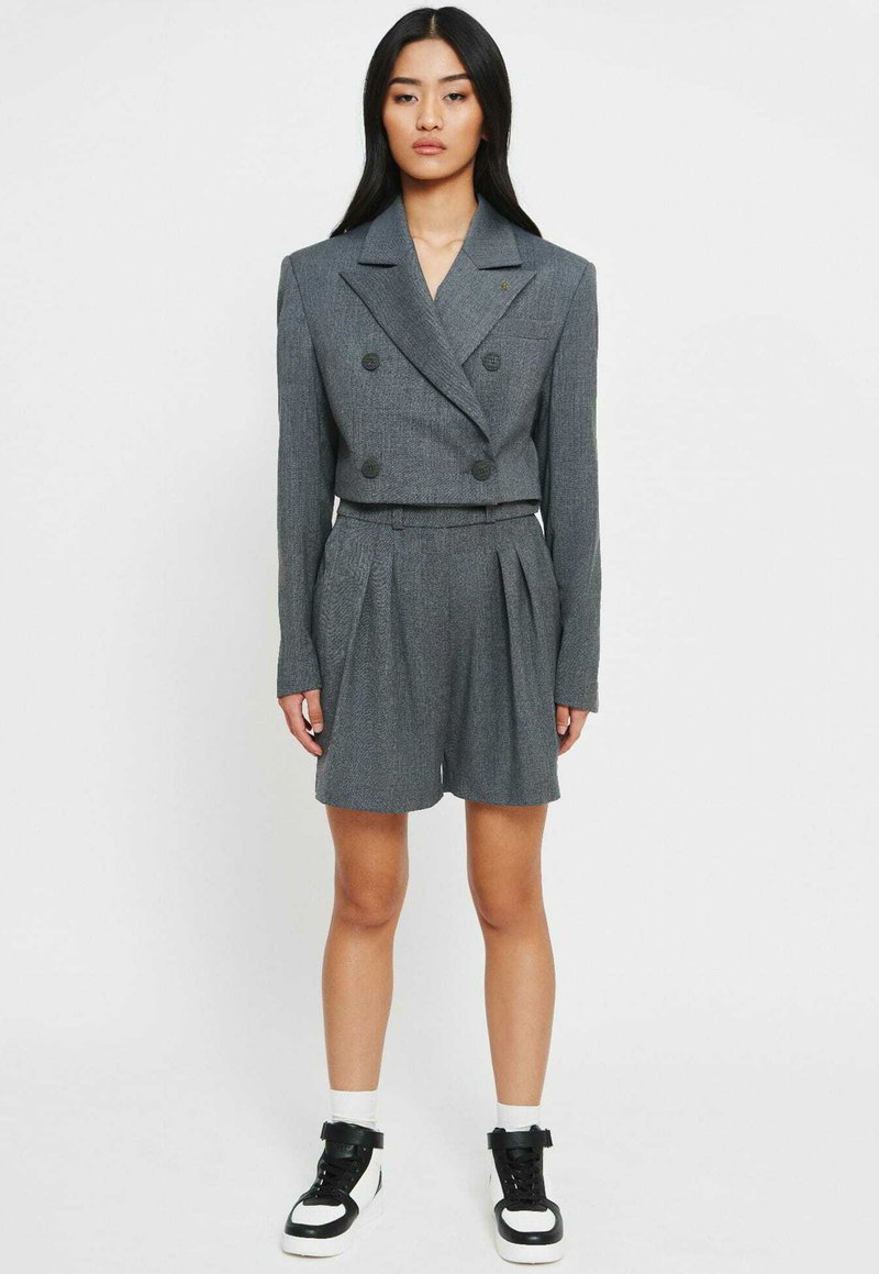 Grijze, dubbelzijdige blazer met schoudervullingen, gecombineerd met bijpassende geplooide short. Heeft een gestructureerde stof en zwarte knopen, gestyled met sneakers.