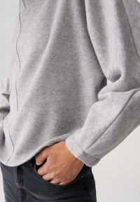 Persona che indossa una maglietta a maniche lunghe grigia chiaro con dettagli cuciti visibili e jeans neri, con una mano posata casualmente nella tasca anteriore.