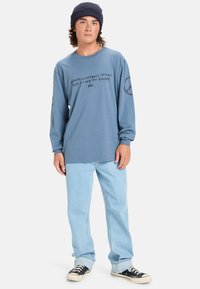 Camisa de manga larga azul con texto negro y diseño de símbolo de paz en las mangas, combinada con vaqueros azul claro y zapatillas negras.