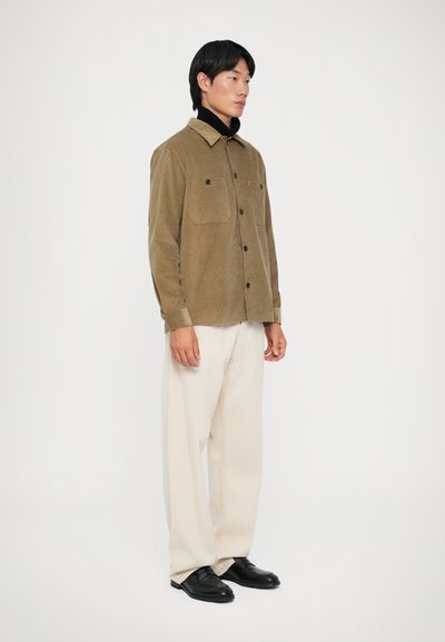 Giacca camicia in velluto a coste marrone con due tasche sul petto, chiusura a bottoni, indossata sopra un dolcevita nero, abbinata a pantaloni a gamba larga color crema e scarpe nere.