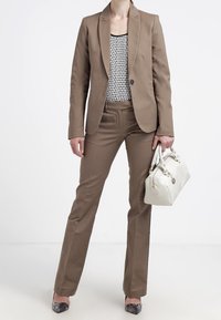 Hnědý upravený blejzr přes vzorovaný černobílý top, ladící hnědé kalhoty, šedé texturované boty a bílá kabelka.