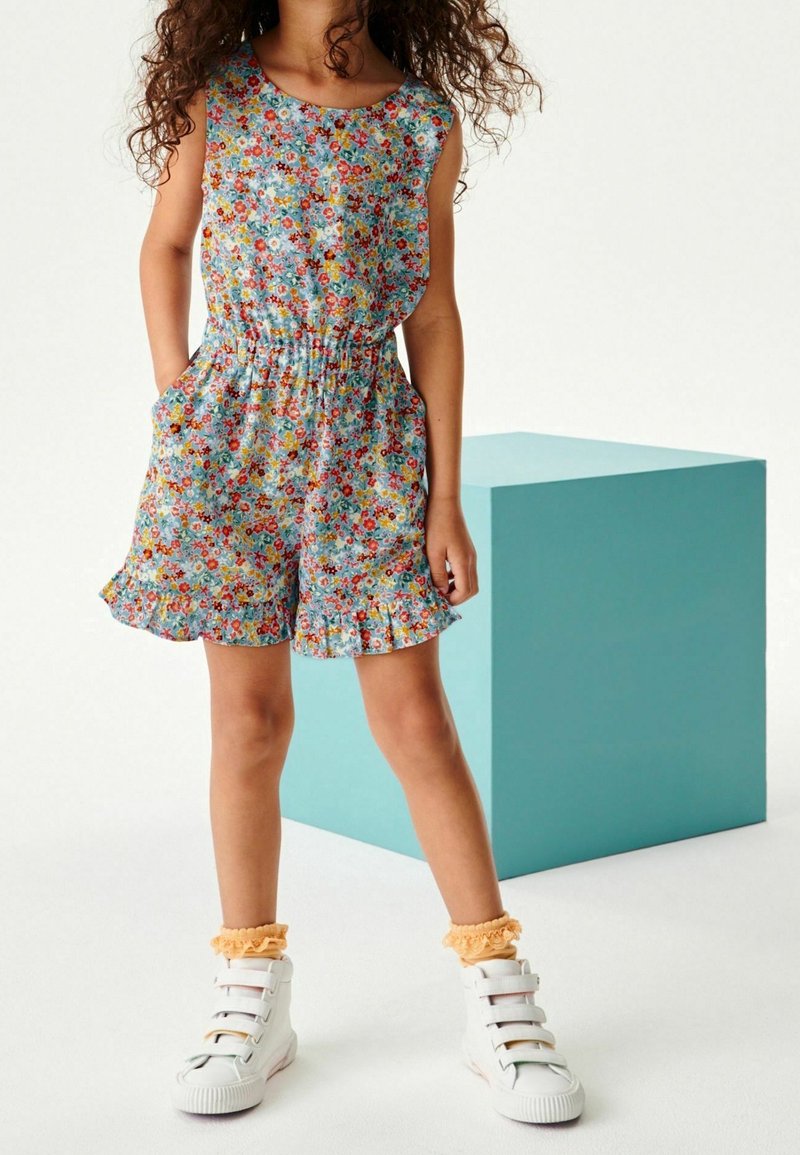 Fille portant une combinaison short sans manches à fleurs avec des shorts à volants, des baskets blanches et des chaussettes chevilles pêche à volants, debout près d'un cube bleu sarcelle.