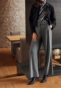 Žena v šedých vysokých kalhotách s širokými nohavicemi, černém sako s knoflíky, vzorovaném roláku a černých špičatých podpatkových kotníkových botách uvnitř.