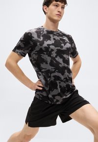 Ανδρικό αθλητικό κοντομάνικο μπλουζάκι σε μαύρο και γκρι καμουφλάζ σχέδιο, κατασκευασμένο από ελαφρύ ύφασμα. Συνδυάζεται με μαύρο σορτς, με άνετη εφαρμογή.