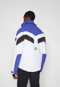 Biela bunda s modrými, čiernymi a sivými prvkami; má vysoký golier, predný zips a logo na chrbte. Text�úrovaný materiál; športový dizajn.