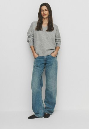 Woman stands against a white background wearing a gray V-neck sweater, wide-leg blue jeans, black shoes, with hands in pockets.