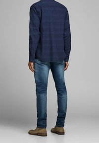 Mörkblå randig långärmad skjorta med knappmanschetter, i kombination med ljusblå slim-fit jeans och tanfärgade mocka ankstövlar. Neutral bakgrund.