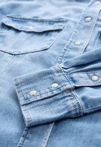 Jasnoniebieska koszula dżinsowa z zatrzaskami, z dwoma kieszeniami na piersi oraz ozdobnym przeszyciem na mankietach i listwie guzikowej.