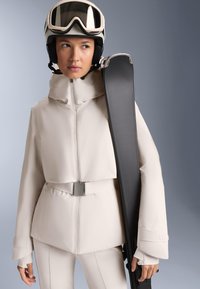 Skijaška odjeća u svijetlo bež materijalu sastoji se od skraćene jakne s remenom, dugim rukavima i uskih hlača koje odgovaraju. Skija je drži sa strane.