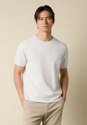 Man staat met handen in de zakken, gekleed in een wit shirt met korte mouwen en beige broek, tegen een effen beige achtergrond.