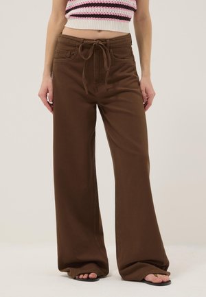 Frau trägt braune, hoch taillierte, weit geschnittene Hose mit Bindegürtel, kombiniert mit schwarzen Flip-Flops und einem kurzen, gestreiften Stricktop.