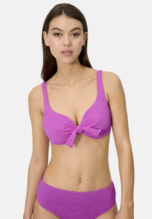 Mujer joven con cabello largo y castaño, vistiendo un bikini morado texturizado con un nudo en la parte delantera, de pie contra un fondo claro y sencillo.