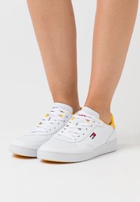 White leather sneakers with yellow accents, round toe, lace closure, and side logo detail. Smooth texture and low-profile design.