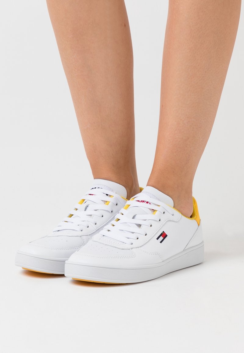 White leather sneakers with yellow accents, round toe, lace closure, and side logo detail. Smooth texture and low-profile design.