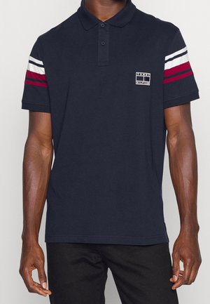 Mann trägt ein dunkelblaues Tommy Hilfiger Polo-Shirt mit weißen und roten Streifen an den Ärmeln und schwarze Hose, steht vor einfarbigem Hintergrund.
