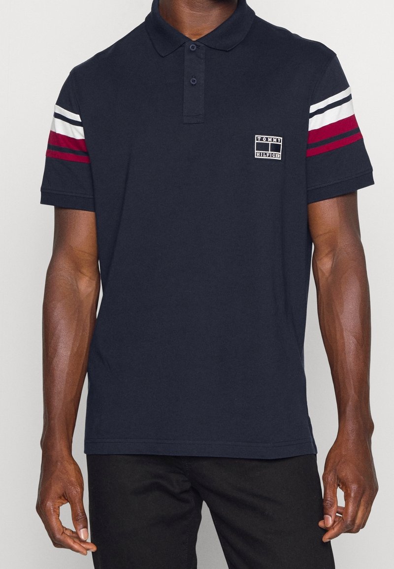 Homme portant un polo bleu marine Tommy Hilfiger avec des rayures blanches et rouges sur les manches et un pantalon noir, debout devant un fond uni.