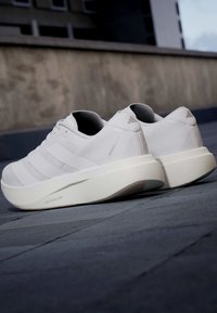Ζευγάρι λευκά αθλητικά παπούτσια Adidas με παχιά σόλα τοποθετημένα σε σκοτεινό πεζοδρόμιο έξω κοντά σε τοίχο κτιρίου.