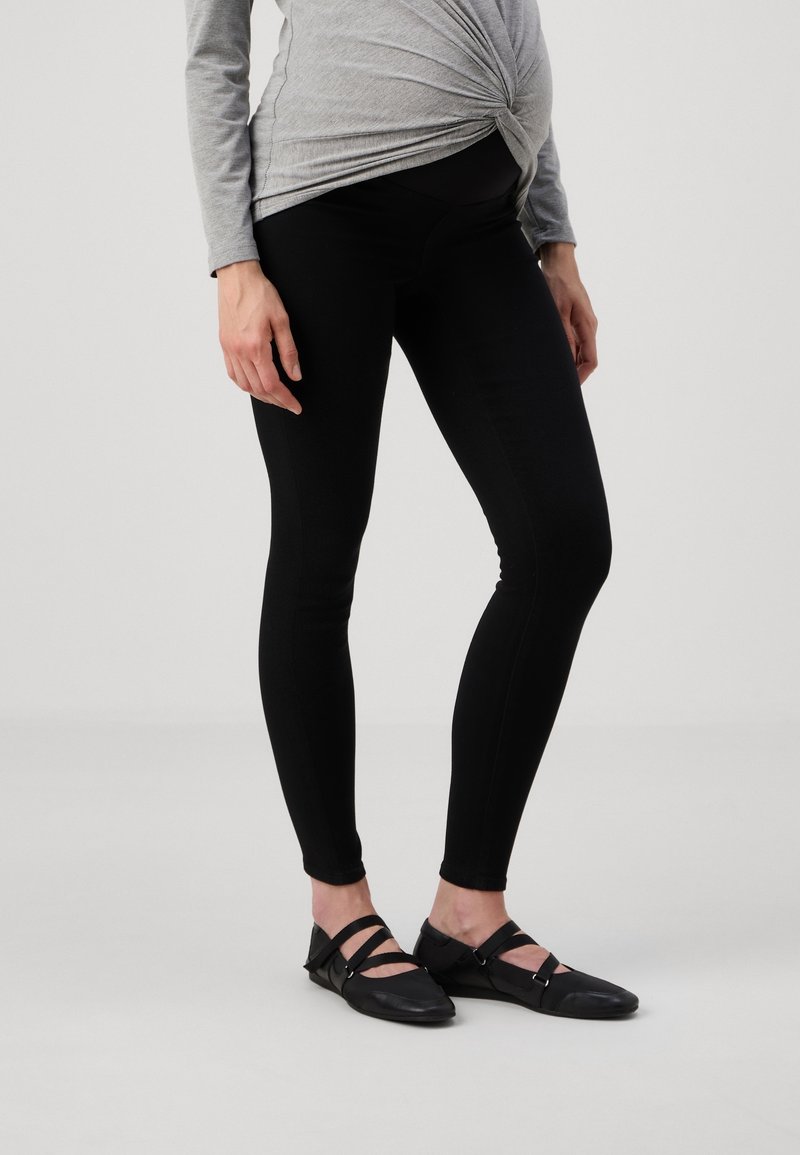 Leggings de maternité noirs en tissu extensible, avec une taille haute et une coupe ajustée. Associés à des chaussures plates noires à brides.
