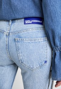 Šviesiai mėlynos džinsinės kelnės su mėlynu žymeniu, ant kurio užrašyta "KARL LAGERFELD JEANS." Ypatybės: dvi galinės kišenės ir mažas logotipo detalė.